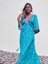Túnica larga de estilo boho, color turquesa con estampado paisley en tonos azules y dorados. Presenta escote en V profundo con bordados decorativos en color fucsia y blanco, mangas amplias tipo kimono y ajuste en la cintura con cordón. El diseño incluye un ruedo con volado y terminaciones con borlas.