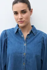 Camisa de jean de color azul medio, con cuello camisero, botones dorados y mangas largas abullonadas con puños ajustados.