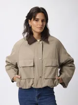 Campera corta de algodón en color beige, con cuello camisero en contraste color marrón, cierre frontal con botones a presión y dos bolsillos frontales tipo cargo.