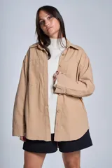 Sobrecamisa color beige confeccionada en gabardina, de corte holgado, con cuello camisero, cierre frontal con botones, dos bolsillos de parche en el pecho y mangas largas con puños abotonados.