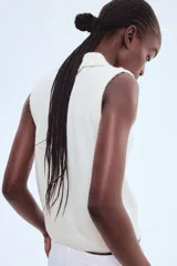 Chaleco corto de punto suave color blanco hueso, con cuello camisero y tapeta de tres botones oscuros. Presenta ribete acanalado en sisas y bajo.