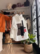 Campera de corderito sintético color blanco, con diseño de collage geométrico en la espalda compuesto por parches en tonos marrón, negro y rosa. Presenta capucha, corte oversize y etiqueta de marca en la parte inferior.