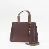 Cartera de mano color bordó con paneles laterales texturizados, manijas superiores en color tostado y dije colgante con cuentas decorativas. Incluye correa larga ajustable y extraíble.