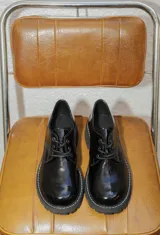 Zapato acordonado de estilo borcego en color negro con acabado charolado, suela track dentada y costuras blancas visibles en el borde de la suela.