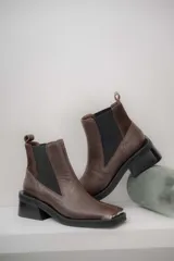 Botas tipo chelsea de cuero color marrón oscuro con acabado brillante. Presentan punta cuadrada con detalle de puntera metálica, elásticos laterales y taco cuadrado bajo.