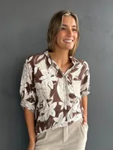 Blusa de corte holgado con estampado floral en tonos marrón y blanco. Presenta cuello camisero, cierre frontal con botones y un bolsillo en el pecho con detalle de lentejuelas. Las mangas son tres cuartos con trabas para ajustar.