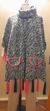 Poncho artesanal gris con cuello alto, bolsillos delanteros con borde rosa y flecos rosas y negros en el ruedo.
