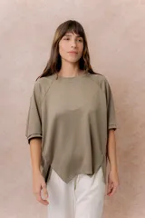 Buzo oversize color beige, con escote redondo, mangas cortas amplias de corte raglán y terminación frontal asimétrica en punta con aberturas laterales.