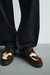 Zapato tipo náutico con diseño de paneles combinados en gamuza marrón y piel con pelo con estampado de vaca. Presenta cordones, horma redonda y suela gruesa con piso track.