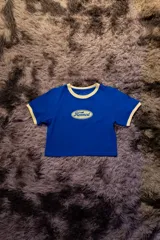 Remera corta de manga corta, color azul vibrante, con cuello redondo y ribetes en color crema. Presenta un logo ovalado centrado con el texto "Hermod" en tipografía cursiva.