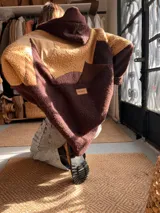 Campera con capucha de diseño patchwork, confeccionada en tejido tipo corderito en tonos marrón y tostado. Presenta un diseño que simula un paisaje montañoso con cortes irregulares y etiqueta de marca en la parte posterior.