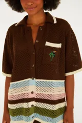 Camisa de punto color marrón con cuello solapa, cierre frontal de botones y mangas cortas. Presenta un bolsillo en el pecho con detalle bordado de una palmera y un tucán, y franjas horizontales multicolor en la parte inferior.
