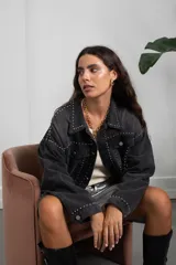 Campera de jean color negro lavado, de corte oversize, con detalle de tachas metálicas plateadas dispuestas en líneas verticales a lo largo de la prenda, cuello clásico, cierre frontal con botones metálicos y bolsillos en el pecho.