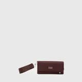 Billetera rectangular de diseño alargado, color bordó, con solapa frontal y logo metálico de la marca. Incluye una correa de muñeca extraíble con el nombre de la marca grabado.