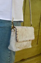 Cartera bandolera de cuero color blanco envejecido con pespuntes marrones. Tiene correa larga regulable y herrajes metálicos color bronce.