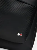 Mochila urbana de diseño minimalista confeccionada en poliuretano negro. Presenta un compartimento principal amplio con cierre, un bolsillo frontal con textura acanalada y el logo de la bandera de la marca en la esquina inferior. Incluye correas de hombro acolchadas y ajustables, además de un asa superior de mano.