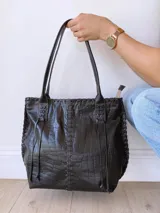 Cartera tipo tote de cuero negro con textura de cocodrilo. Presenta detalles de costuras trenzadas a lo largo de la estructura y tiras decorativas laterales. Cuenta con doble asa de hombro y cierre superior.