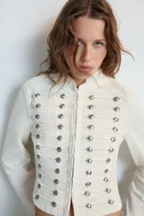 Chaqueta de denim blanca con cuello redondo, manga larga y corte entallado. Presenta un diseño de inspiración militar con múltiples tiras horizontales en el delantero decoradas con botones metálicos plateados, y cierre frontal con ganchos.