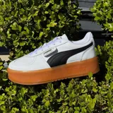 Championes Puma modelo Palermo, con diseño clásico de perfil bajo. Presentan una combinación de cuero blanco y detalles en gamuza gris claro, con la icónica franja lateral de Puma en color negro. Incluyen suela de goma color caramelo y detalles de la marca en dorado.