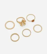 Kit de seis anillos de metal en tono dorado con diversos diseños, incluyendo un anillo con detalle de lazo, uno con piedra pequeña, uno trenzado, uno texturizado y dos lisos.