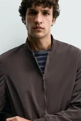 Cazadora bomber ligera de tejido técnico color verde oliva, con cuello, puños y bajo en rib elástico. Presenta cierre frontal de cremallera y bolsillos de vivo en la cadera.