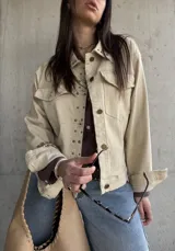 Sobrecamisa beige de gabardina con tachas metálicas decorativas en el cuello y la solapa frontal. Cuenta con bolsillos con solapa en el pecho y cierre frontal con botones.