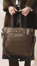 Cartera tipo tote marrón de diseño práctico y versátil, confeccionada en símil cuero con textura granulada. Cuenta con asas cortas fijas, bolsillos exteriores con cierre al frente y atrás, y bolsillos laterales abiertos. Su interior es amplio e incluye bolsillos organizadores. Viene con una correa ajustable y desmontable.
