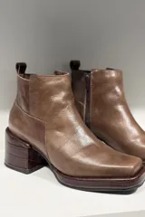 Bota corta de cuero color marrón, con diseño de punta cuadrada y taco cuadrado con textura grabada. Presenta costuras visibles y una lengüeta en la parte posterior.