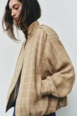 Chaqueta bomber de color beige con diseño a cuadros blancos, cuello subido, manga larga con trabillas en los hombros, bolsillos delanteros y bajo con elástico. Cierre frontal con cremallera y botones a presión.