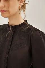 Camisa blanca de algodón con cuello alto, cierre frontal de botones ocultos y detalles de bordado floral en el pecho. Presenta mangas largas con puños elásticos y volados decorativos en la parte frontal.