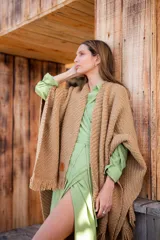 Ruana tejida en color camel con textura de punto grueso y terminaciones con flecos en los bordes. Presenta una etiqueta de cuero sintético en la parte inferior.