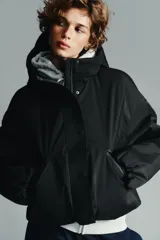 Anorak corto de color negro con acolchado interior, cuello subido y capucha. Presenta mangas largas con puños elásticos, bolsillos delanteros con cierre y bajo ajustable con cordones. Cierre frontal con cremallera termosellada y botones a presión.