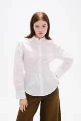 Camisa blanca de poliéster con cuello clásico, manga larga con puños anchos y detalle de frunces en la cintura que crea un efecto entallado.