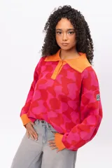 Buzo de tejido de punto con diseño geométrico en tonos fucsia y rojo, cuello tipo polo con cierre de botones y puños en color naranja contrastante. Presenta un corte holgado y oversize.