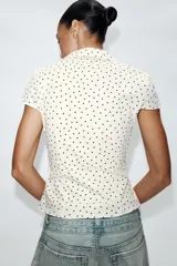 Camisa de cuello solapa y escote en pico, con mangas cortas y detalle de frunces centrales. Presenta cierre frontal con botones y estampado de lunares negros sobre fondo blanco.