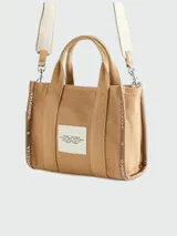 Cartera tipo tote bag de lona color beige, con diseño estructurado y asas superiores. Presenta el logo de la marca estampado en la parte frontal y detalles de cintas laterales con inscripciones.