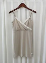 Camisón maternal color beige, con diseño de escote cruzado y breteles finos ajustables. Presenta detalles de puntilla blanca en el escote y un pequeño aplique de marca en el ruedo.