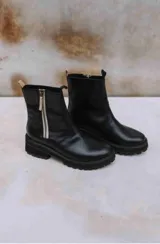 Botas cortas de cuero negro con suela de goma gruesa y dentada. Presentan un cierre lateral con cremallera expuesta de color claro (posiblemente blanco o beige) y tiradores de cuero en la parte superior.