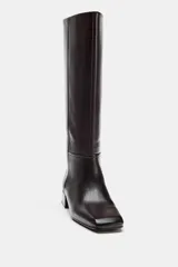 Bota de piel color marrón oscuro, de caña alta, con detalle de pespuntes en el cuerpo, tacón medio en bloque y punta cuadrada.