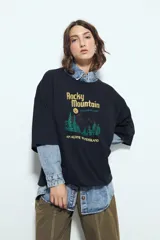 Remera blanca de manga corta con calce relajado y cuello a la base. Presenta un estampado frontal pequeño y una estampa protagonista en la espalda con el texto "Rocky Mountain National Park" y un paisaje de montañas y árboles.