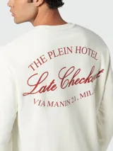 Buzo de cuello redondo color crema o blanco roto, de manga larga y corte regular. Presenta un estampado frontal en color rojo oscuro que simula un escudo heráldico con el texto "The PLEIN HOTEL MILAN".