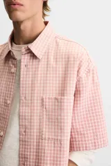 Camisa de corte relajado confeccionada en algodón, con estampado de cuadros pequeños en tonos rosados. Presenta cuello solapa, manga corta y bolsillo plastrón en el pecho con cierre frontal de botones.