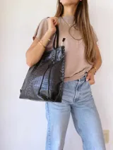 Cartera tipo tote de cuero negro con textura de cocodrilo. Presenta detalles de costuras trenzadas a lo largo de la estructura y tiras decorativas laterales. Cuenta con doble asa de hombro y cierre superior.