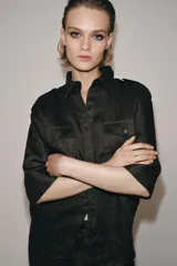 Camisa de lino color marrón oscuro con cuello solapa, manga por debajo del codo y cierre frontal con botones ocultos por solapa. Presenta bolsillos de plastrón con solapa en el pecho y trabillas en los hombros.