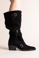 Bota alta de gamuza color negro, sin forro y con caída natural en la caña ancha. Cuenta con taco cuadrado, punta fina y tres tiras ajustables con hebillas metálicas plateadas.