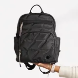 Mochila negra acolchada con cierre superior y bolsillo frontal con cierre. Tiene manija superior y correas ajustables para los hombros. Incluye un dije decorativo de cuerdas trenzadas multicolores.
