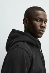 Sudadera con capucha y cierre de cremallera, color negro, de corte relajado y tejido de algodón compacto y pesado con interior rizo. Posee bolsillos frontales tipo canguro y acabados en rib.