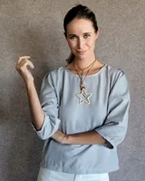 Blusa de crepe color gris claro, con mangas tres cuartos que presentan cortes y pespuntes decorativos. Posee un diseño holgado y cuello redondo.