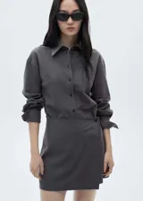 Vestido corto de estilo camisero con diseño cruzado, cuello clásico y cierre frontal de botones. Presenta mangas largas con puños abotonados y un sutil estampado de raya diplomática.
