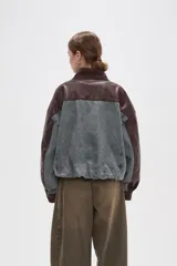 Campera estilo bomber con diseño color block, combinando paneles de denim lavado con mangas y detalles en cuero sintético color marrón oscuro. Presenta cuello alto con tira ajustable, cierre frontal con botones a presión y bolsillos frontales tipo parche.